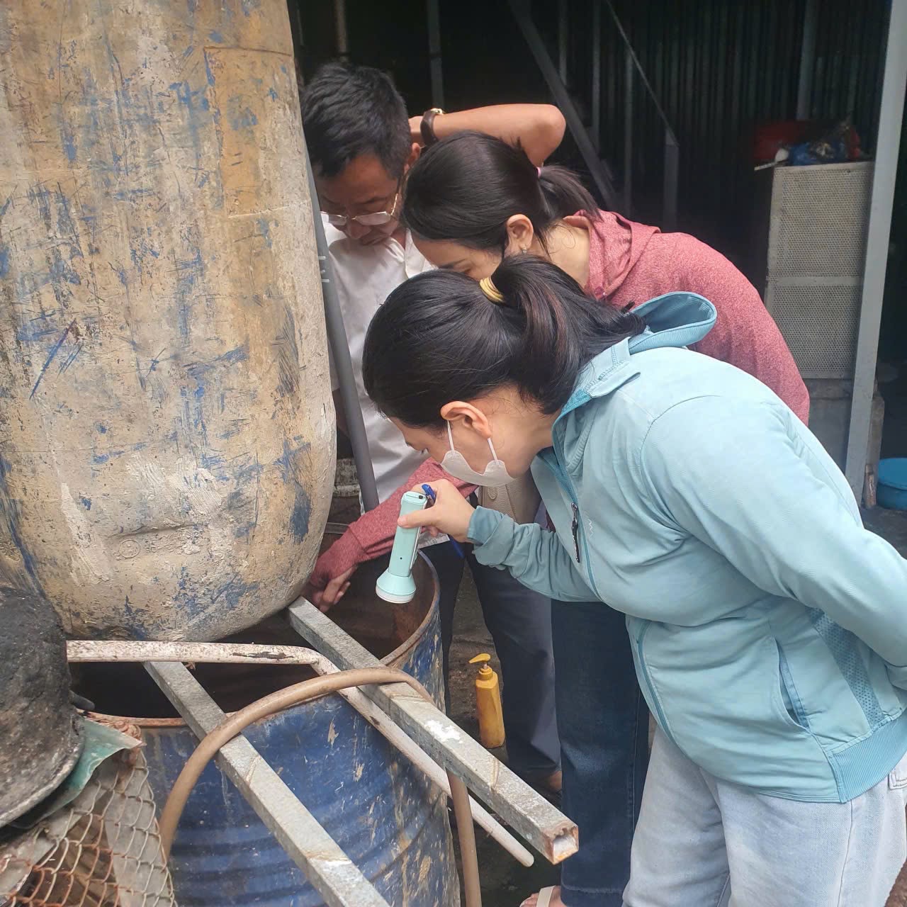 Cán bộ y tế dùng đèn chiếu kiểm tra kỹ lưỡng các thùng phuy chứa nước kim loại tại khu vực sản xuất.